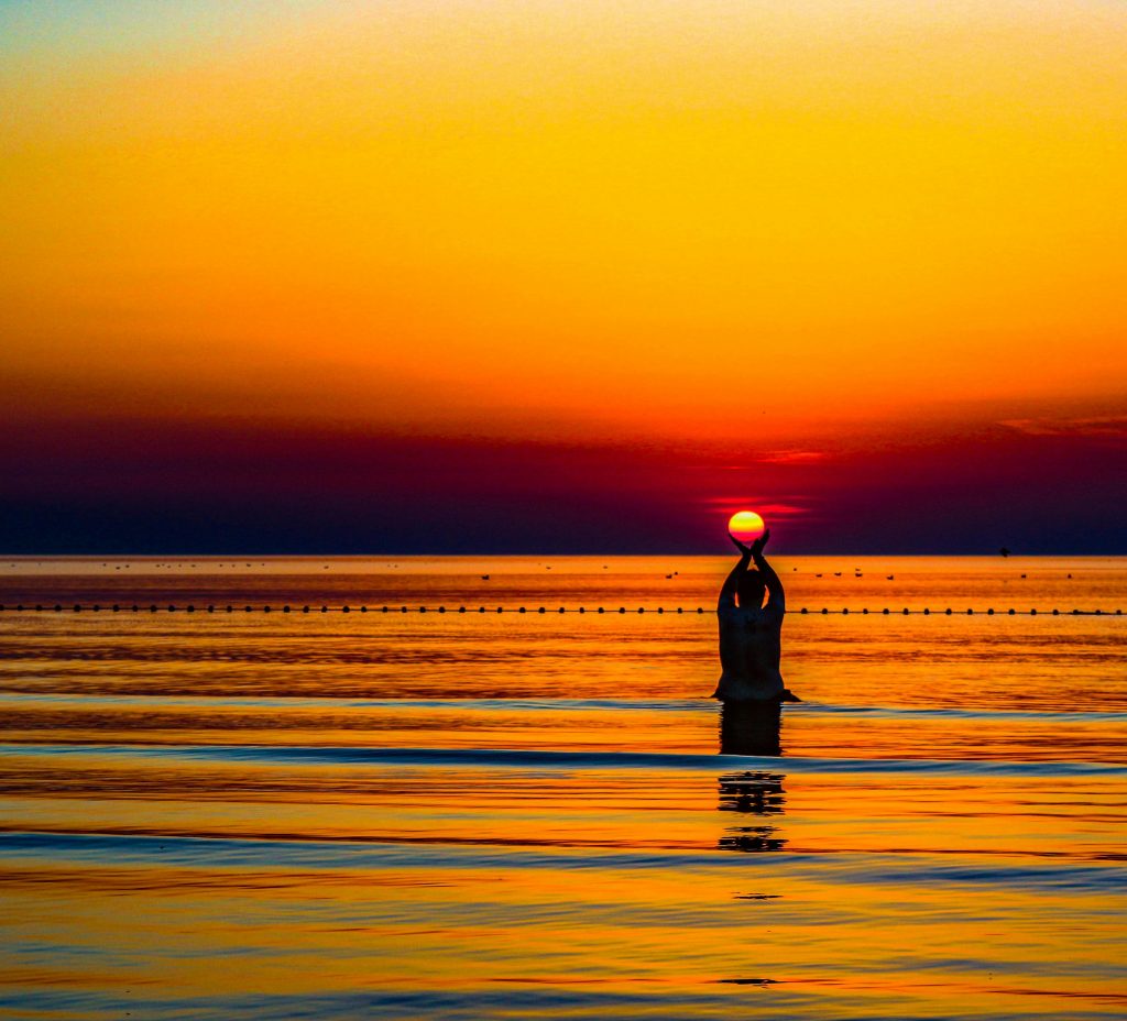 A serene silhouette of a person holding the sun at sunset over a calm sea, capturing tranquility.